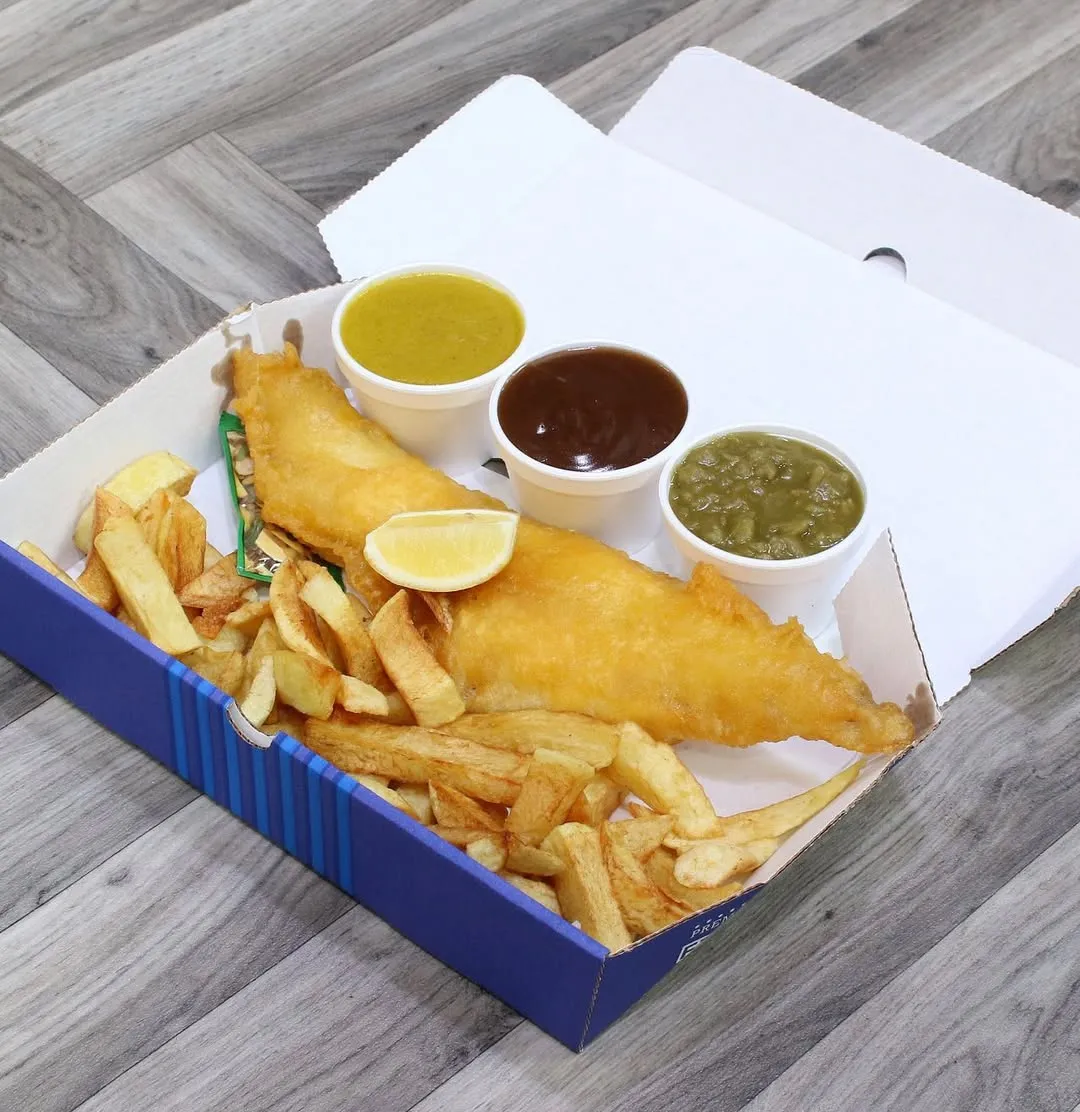 The image showcases a well-composed and brightly lit serving of fish and chips in a takeaway box, accompanied by three distinct sauces and a lemon wedge. The food items are sharp and appear photo-realistic with accurate, appealing colors. All key elements are in clear focus, making the dish look appetizing and ready to eat. The overall presentation is professional, emphasizing the quality of the meal.
