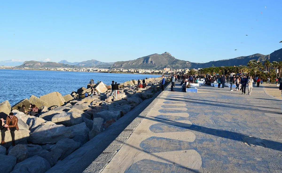 The image is a well-lit and sharp photograph of a coastal scene, showcasing a promenade with people along a rocky shore and mountains in the background. The colors are natural and accurate, contributing to a high degree of photo-realism. Key elements from the foreground pathway to the distant mountains are in good focus. The composition is generally effective, although the foreground pathway occupies a substantial portion of the frame.