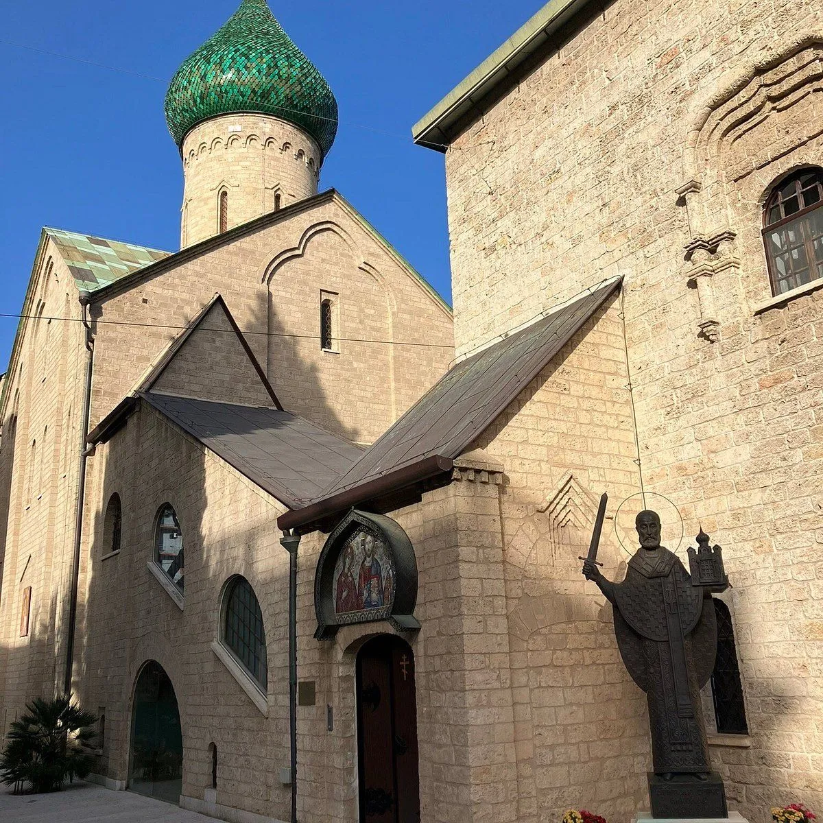 An exterior view of a historical stone building featuring a distinctive green-tiled dome, set against a clear blue sky. A statue of a figure holding a sword and a miniature building stands prominently to the right.
