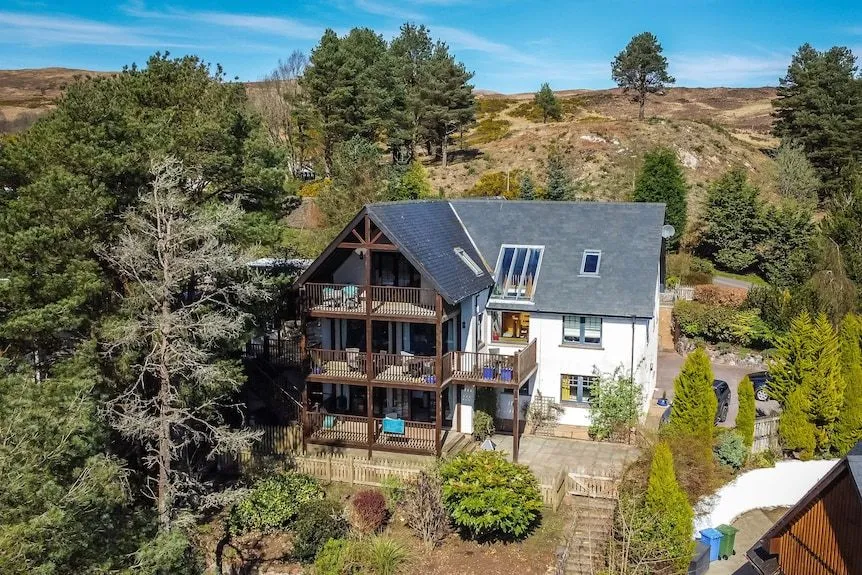 An aerial view of a large, multi-story house with several wooden balconies, set amidst green trees and rolling hills under a clear blue sky.