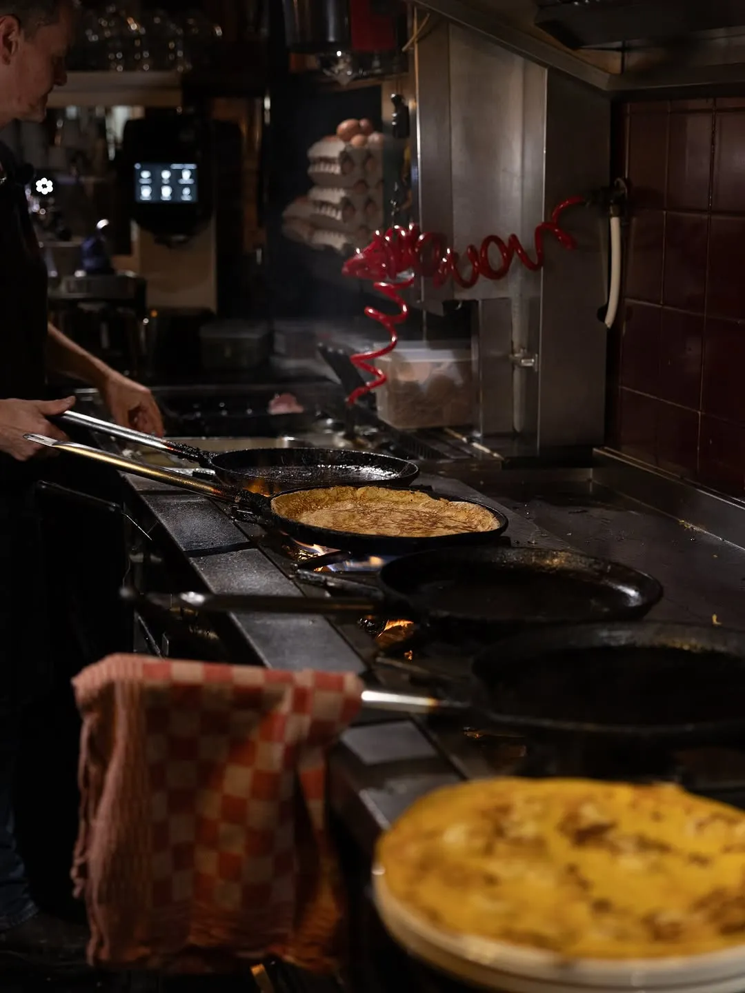 The image effectively captures a dynamic kitchen scene, highlighting the process of making crepes or pancakes. It is photo-realistic and the colors appear accurate for the environment. Key elements, such as the food in the pans, are in focus, contributing to the visual appeal. However, the overall lighting is somewhat dim, and while the composition is good, some areas of the image lack optimal sharpness.