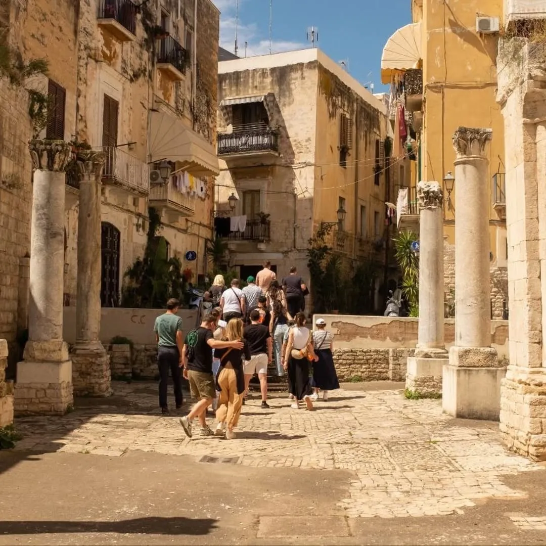 An old town street scene with ancient pillars, historical buildings, and people walking up an incline under bright daylight.
