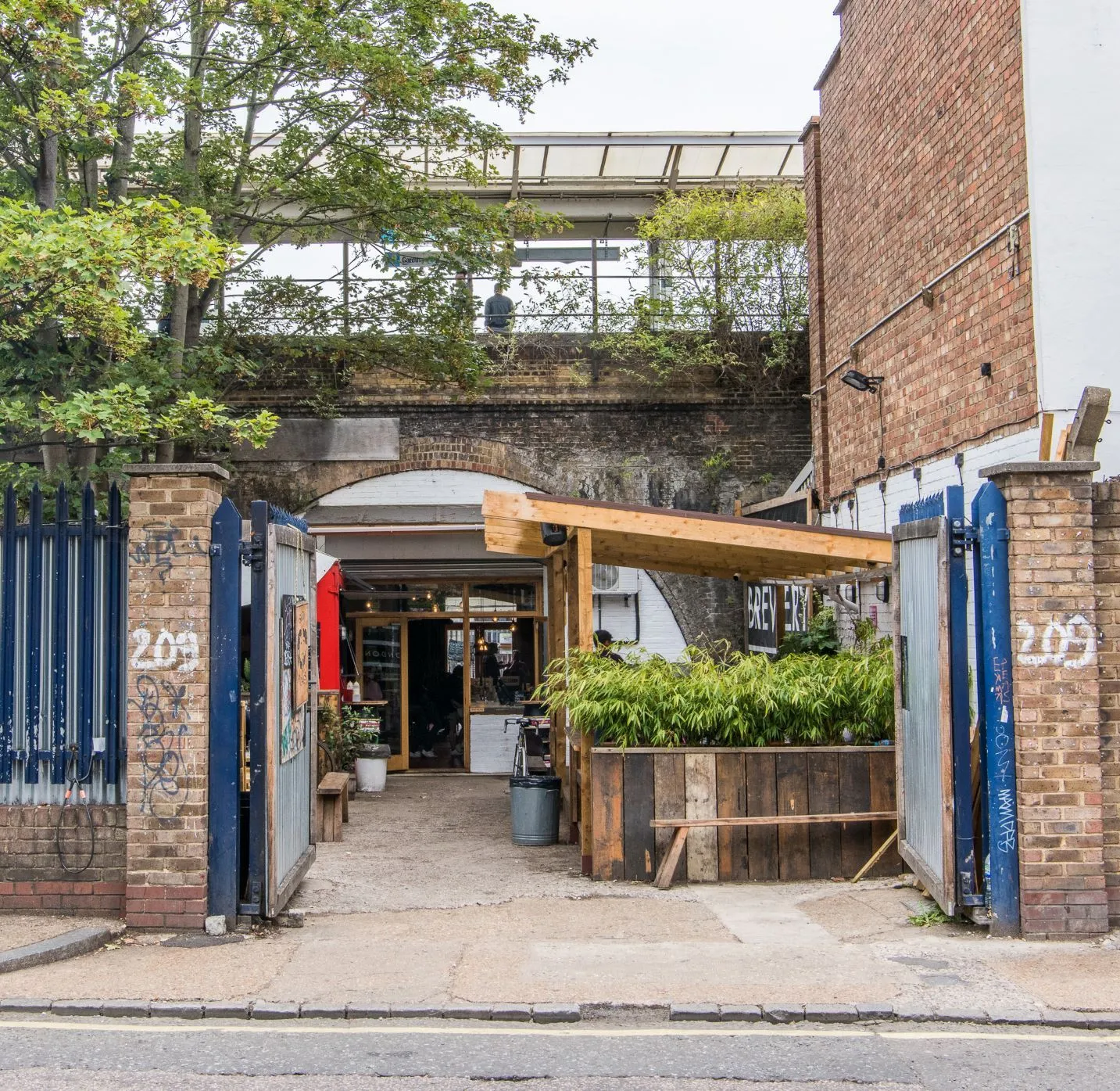 An exterior shot of a brewery entrance located under a railway arch. The image displays good sharpness and natural, even lighting, resulting in accurate colors and a high degree of photo-realism. Key elements, such as the entrance and surrounding structures, are in clear focus. The composition is generally well-balanced, though the cropping of the trees at the top could be slightly improved.