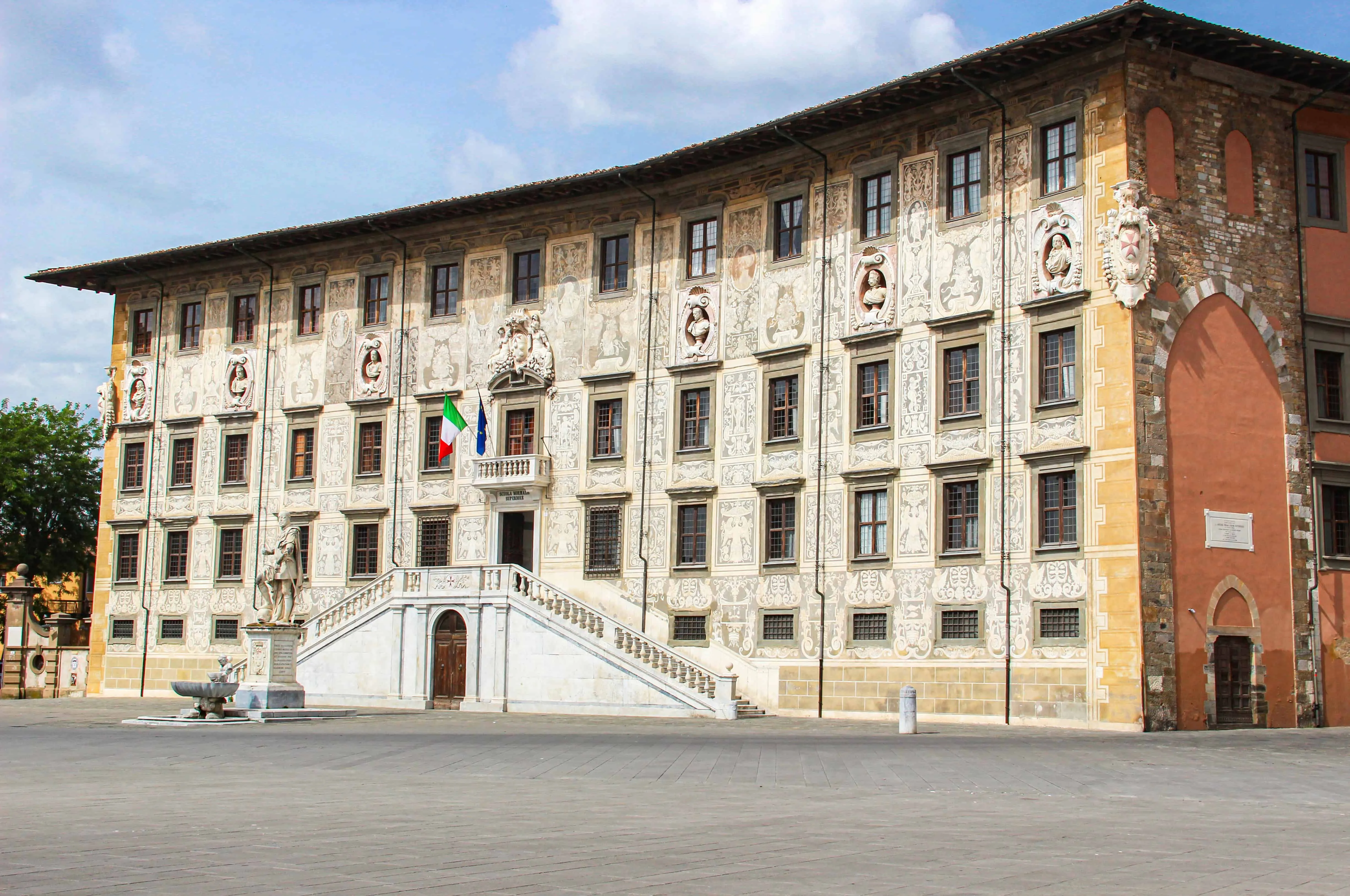 An editorial-style image of a grand, historic building with intricate facade details and a prominent grand staircase, set under a clear blue sky. Italian flags are visible near the entrance.