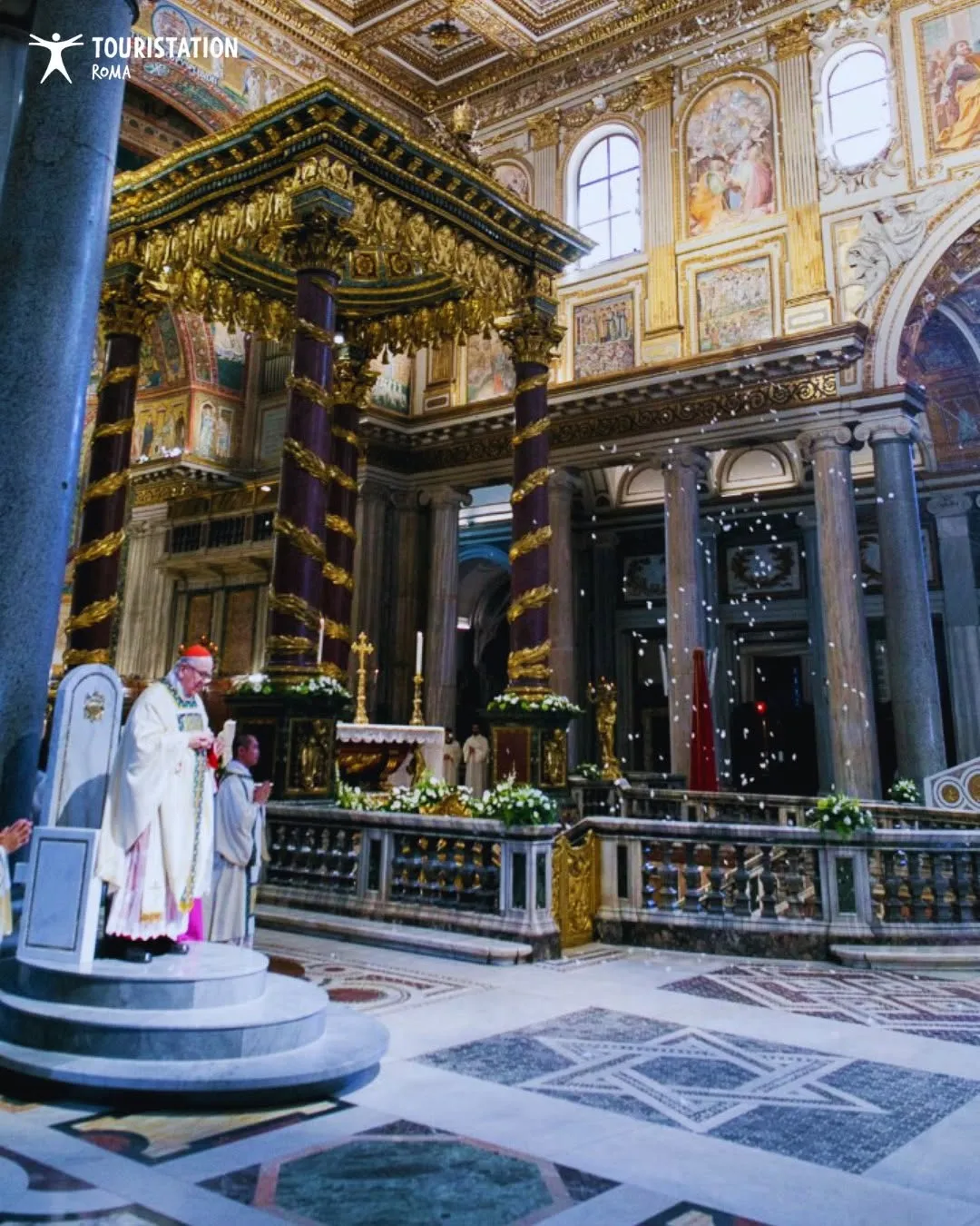 An indoor image capturing a religious ceremony within a grand, ornate church. A cardinal or priest is visible in the foreground, standing on a raised platform, with elaborate altar structures and columns filling the background. Small white specks appear to be falling through the air.