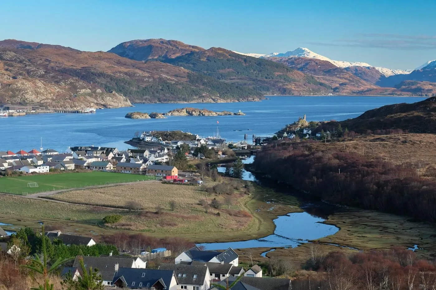 An expansive view of a Scottish coastal village nestled beside a body of water, with mountains in the background under a clear blue sky.
