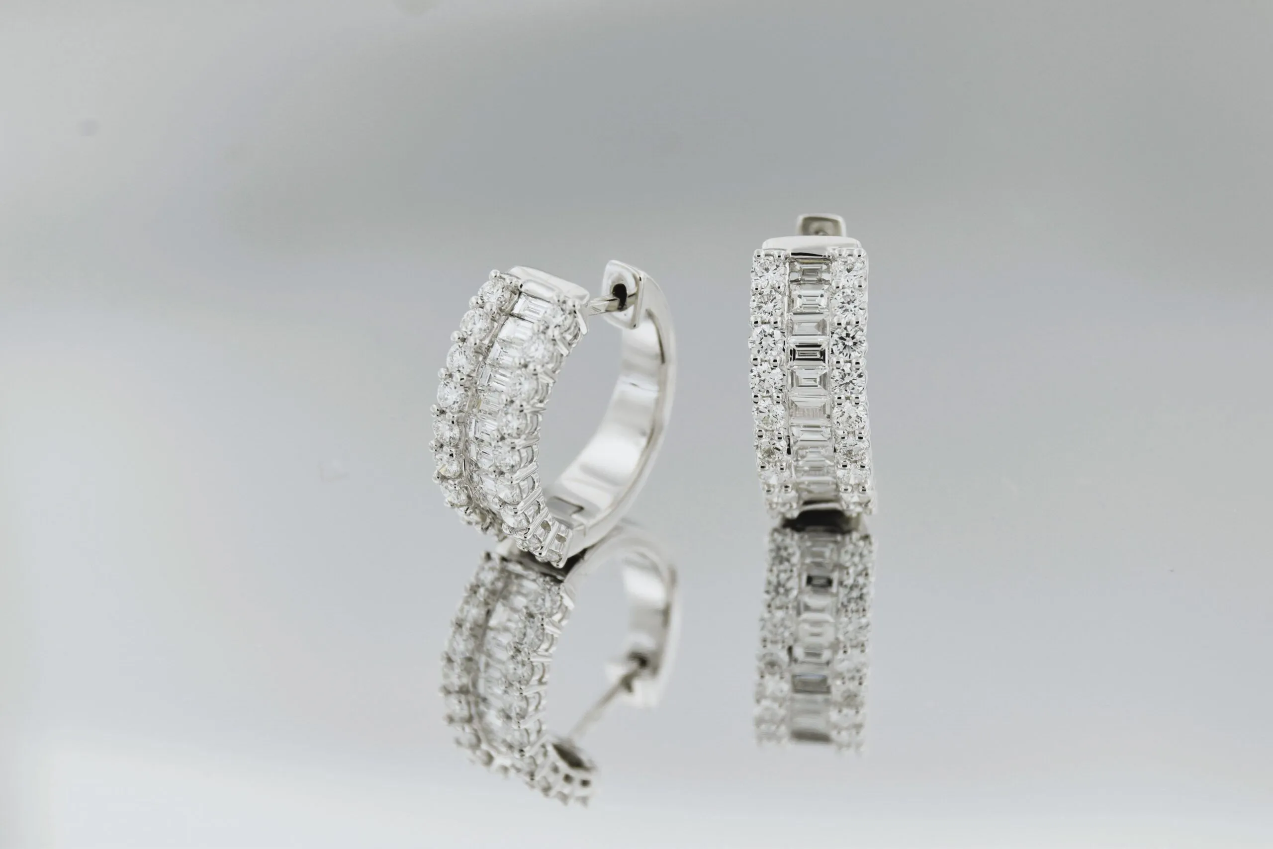 A pair of white gold or platinum diamond hoop earrings, one slightly in front of the other, displayed on a reflective surface with a neutral gray background.