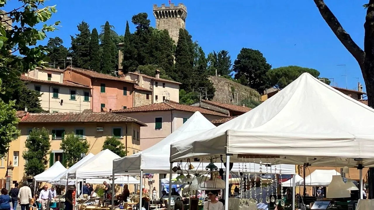 An outdoor market is pictured in the foreground, with several white tents housing vendors. In the background, traditional Italian buildings with terracotta roofs and green shutters ascend a hill, crowned by a historic stone tower. The scene is bathed in bright daylight under a clear blue sky.