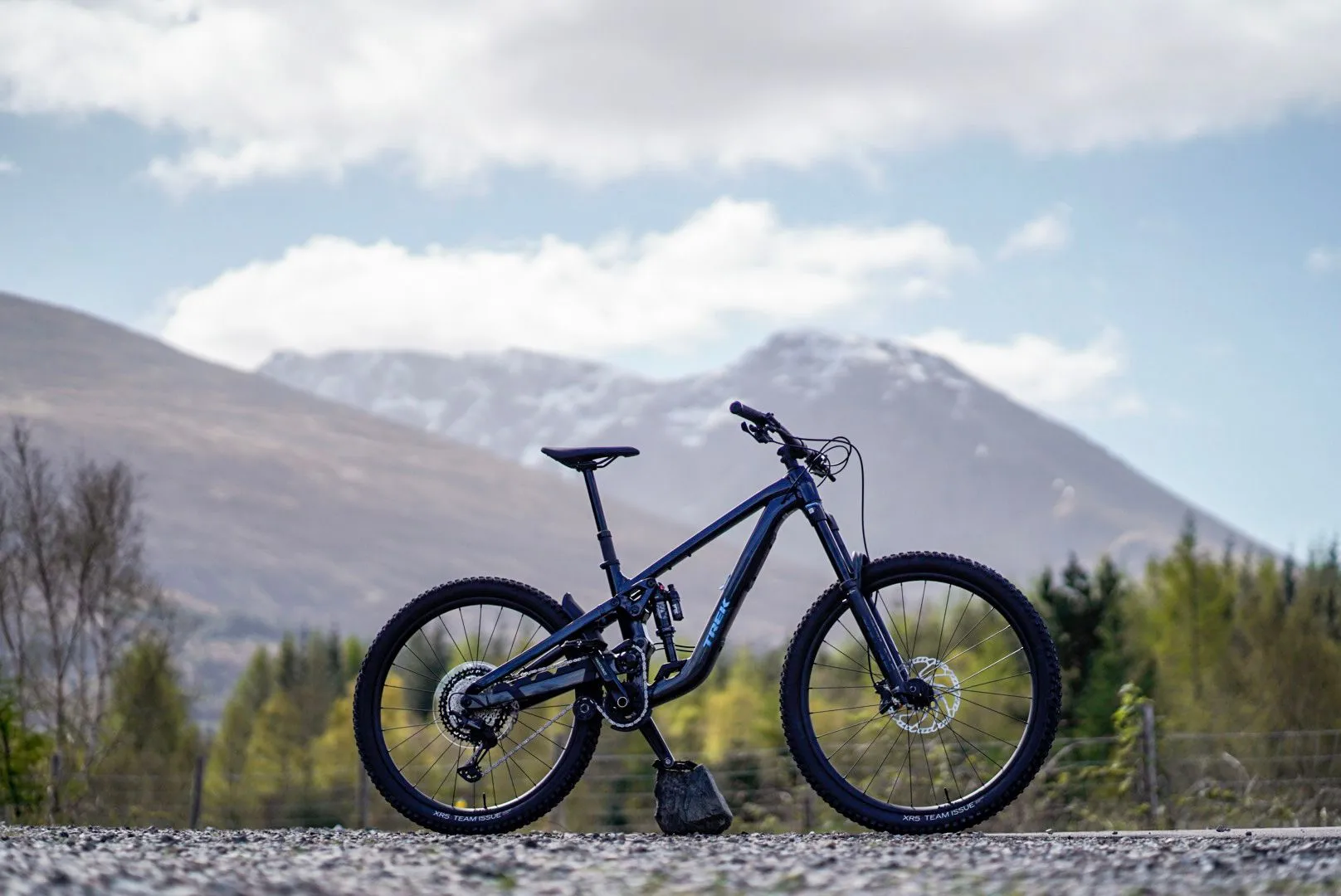 A high-quality photograph of a dark-colored mountain bike standing on a gravel surface, set against a backdrop of mountains and a partly cloudy sky. The bike is the central focus, sharply defined.