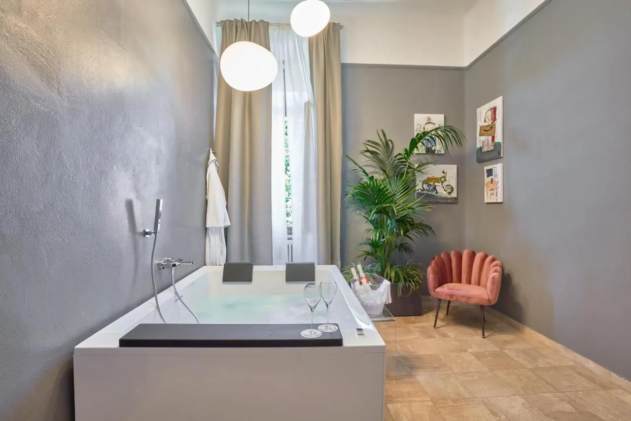 An editorial style image showcasing a luxurious and modern bathroom space with a large bathtub, two champagne flutes, a bottle of champagne in an ice bucket, a plant, a decorative chair, and framed artworks on the wall. The room features grey walls and light-colored curtains.