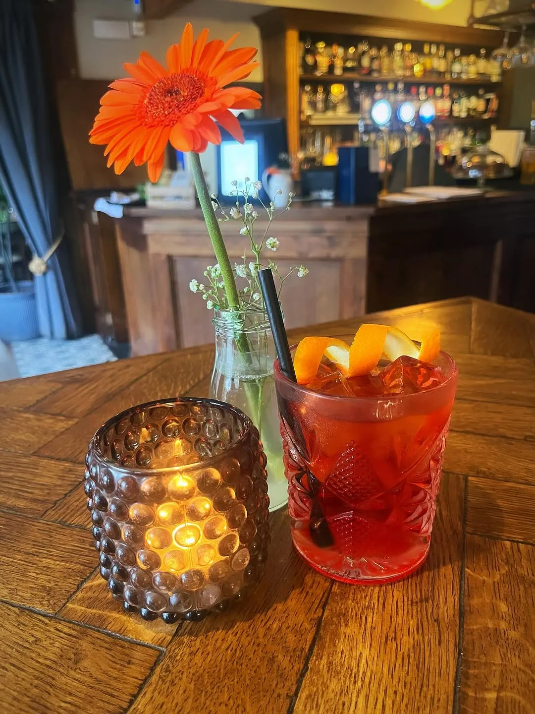 A well-composed image featuring a vibrant cocktail, an orange flower, and a lit candle on a wooden table. The primary subjects are in focus, with a pleasingly blurred background of a bar, contributing to a photo-realistic and atmospheric shot. The colors are generally accurate and appealing.