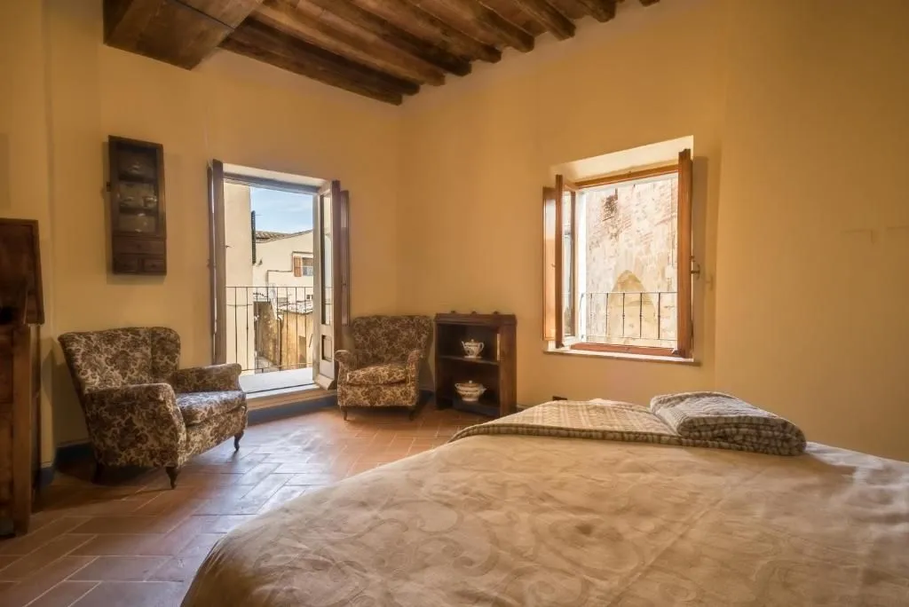 An editorial-style image of a bedroom featuring traditional decor. The room includes a large bed in the foreground, two patterned armchairs, and a small wooden bookshelf. Two open windows, one leading to a balcony, offer views of an old town or city outside. The room features terracotta tile flooring and wooden ceiling beams.