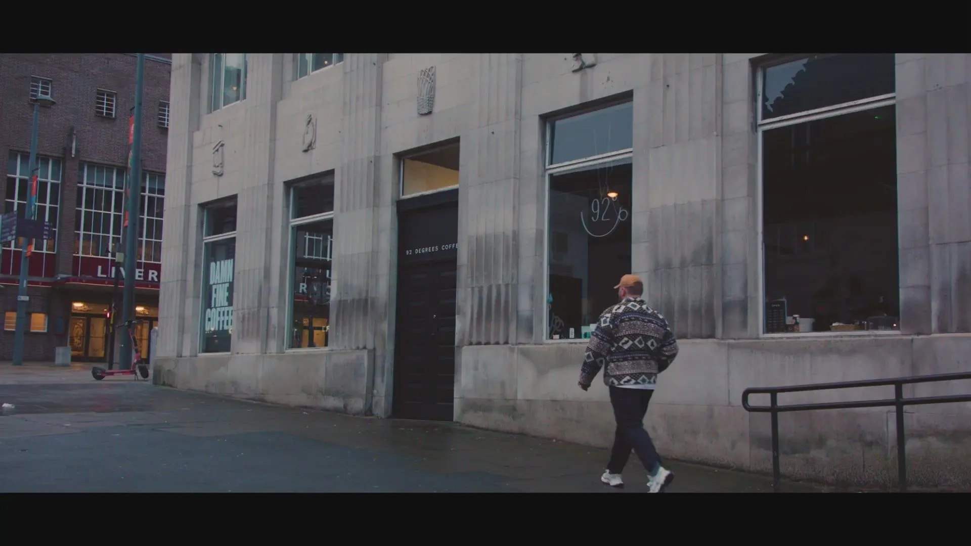 The image features a well-composed street scene with a person walking past a coffee shop. It is photo-realistic, and key elements like the person and the building are in focus. The natural, somewhat overcast lighting is accurate, contributing to an authentic urban aesthetic, though it limits overall vibrancy. The sharpness is good for the scene depicted.