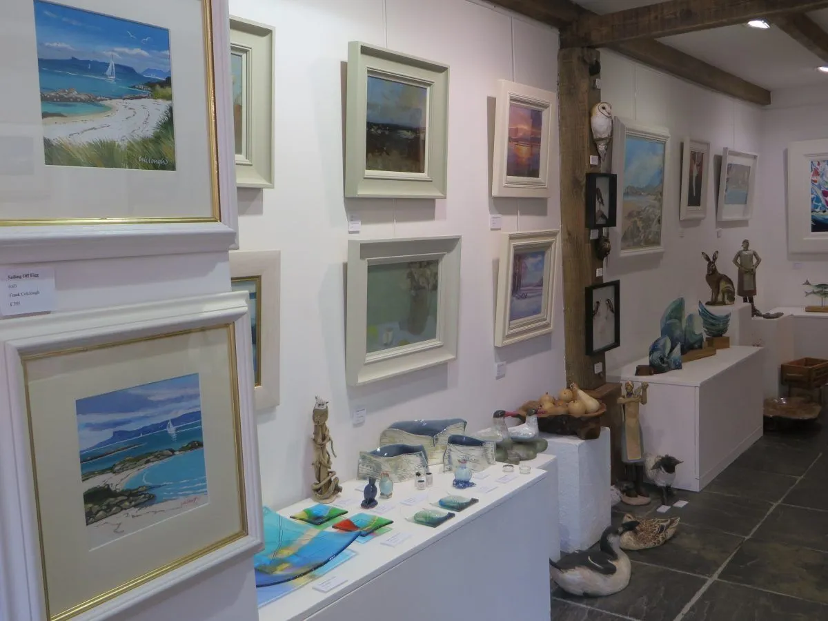 The image captures an art gallery interior, showcasing various paintings hung on white walls and sculptures displayed on pedestals. The space appears well-lit, highlighting the artworks.