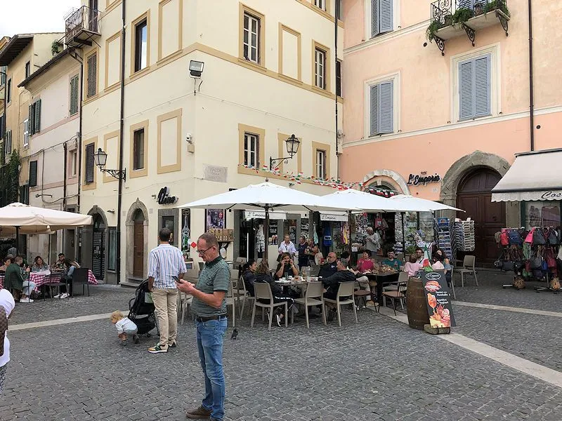 An authentic street scene depicting an outdoor cafe in a European town square, featuring historical buildings and various people, some dining and others walking.