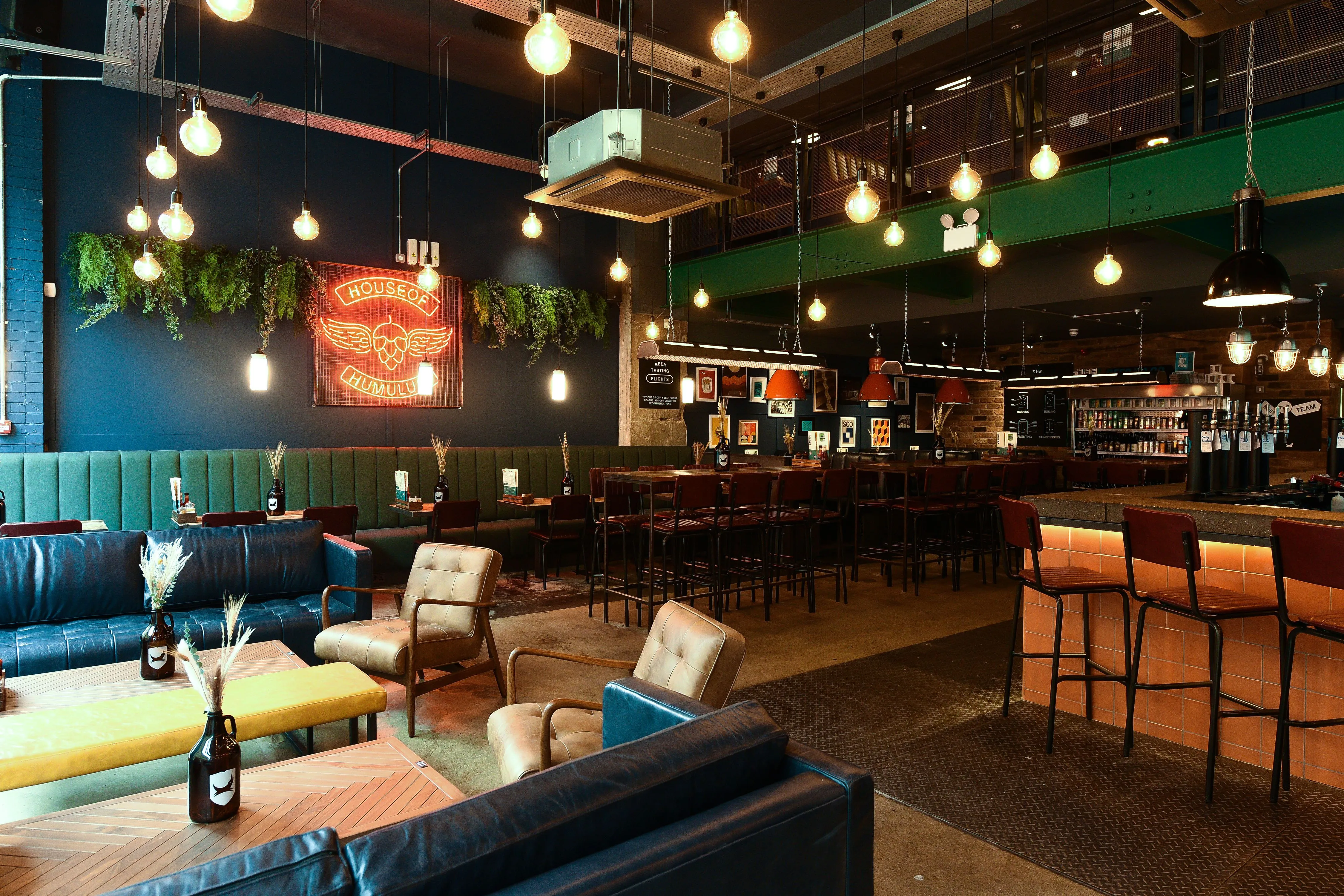 An inviting, dimly lit bar or restaurant interior with a mix of industrial and cozy elements, featuring various seating arrangements, a prominent neon sign, and atmospheric lighting.