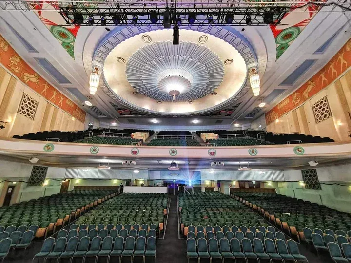An interior wide shot of an empty grand theater or auditorium, featuring rows of green seats, an ornate ceiling with circular patterns, and a stage area illuminated by a projector beam.