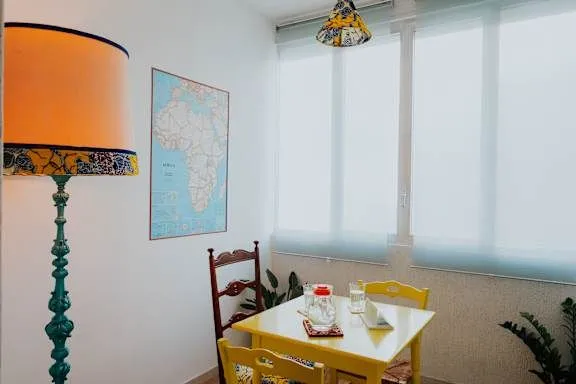 A bright and inviting dining space featuring a vibrant yellow square table and two matching yellow chairs. A third, darker wooden chair is also visible. On the wall, a framed map of Africa adds a decorative touch. Natural light streams in from large windows, creating a clean and domestic atmosphere.