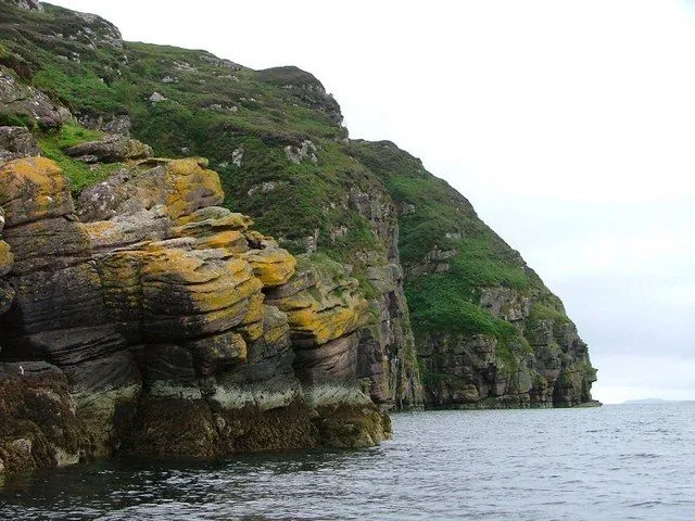 The image is an editorial quality shot of a rugged coastline, featuring impressive cliffs covered in green vegetation and textured rocks meeting the sea. The composition effectively showcases the natural elements.