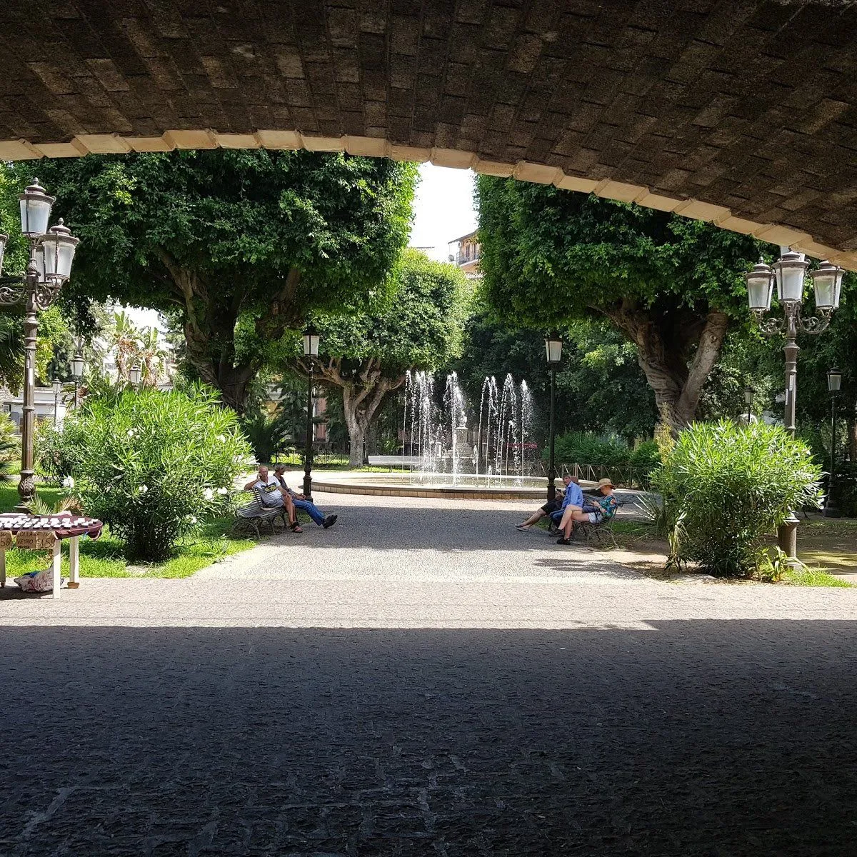 The image features a photo-realistic scene of a park with a central fountain, framed by a stone archway. Strengths include good color accuracy, sharp focus on key mid-ground elements like the fountain and trees, and a generally well-composed frame utilizing the archway. The image captures a natural, everyday scene. The primary weakness is the significant contrast in lighting, with the foreground under the archway being very dark and lacking detail compared to the well-lit park in the background.