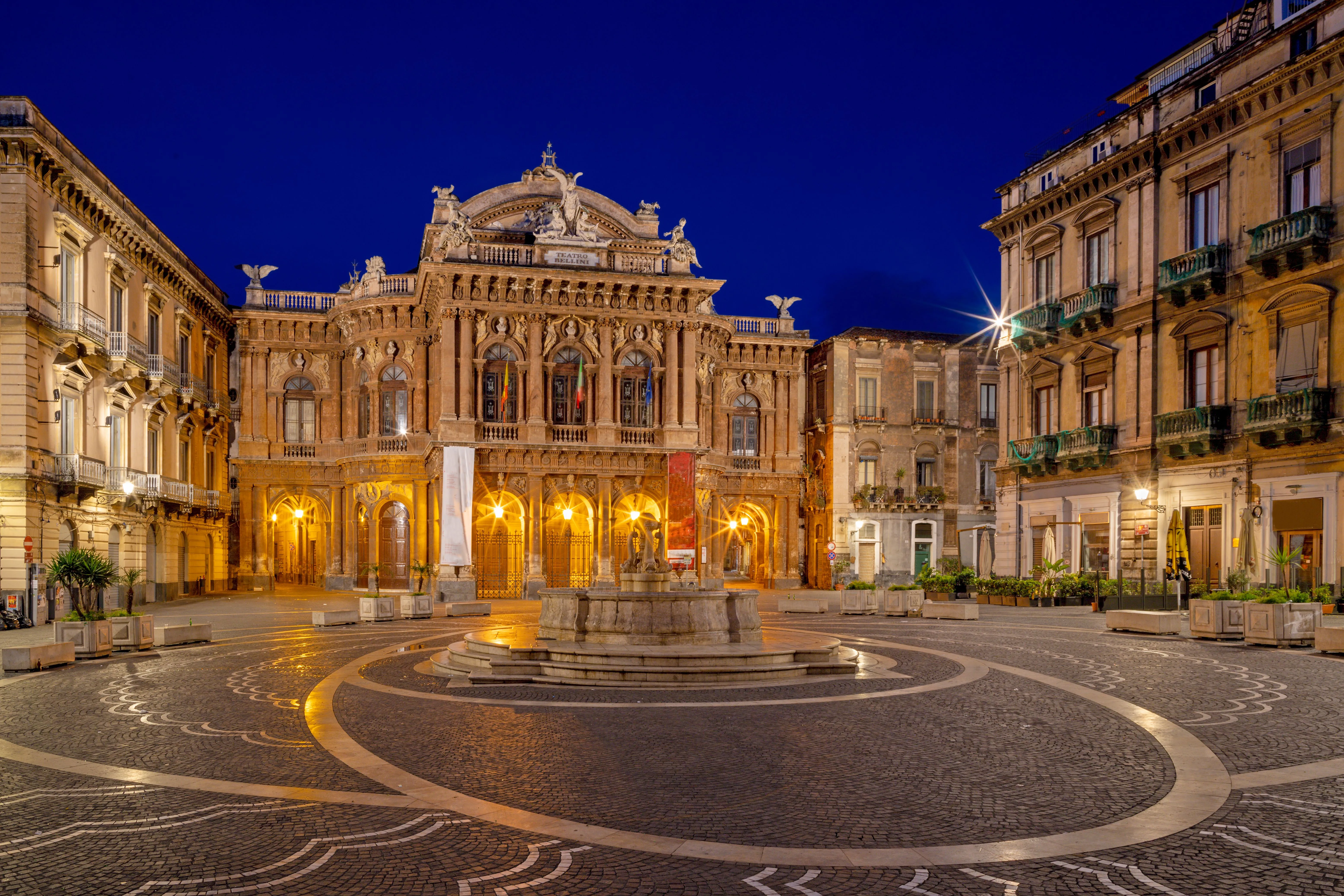 An exceptionally well-composed photograph of a grand, ornate building and a fountain in a city square at night. The deep blue sky provides a striking contrast to the warm, golden light illuminating the architectural details.