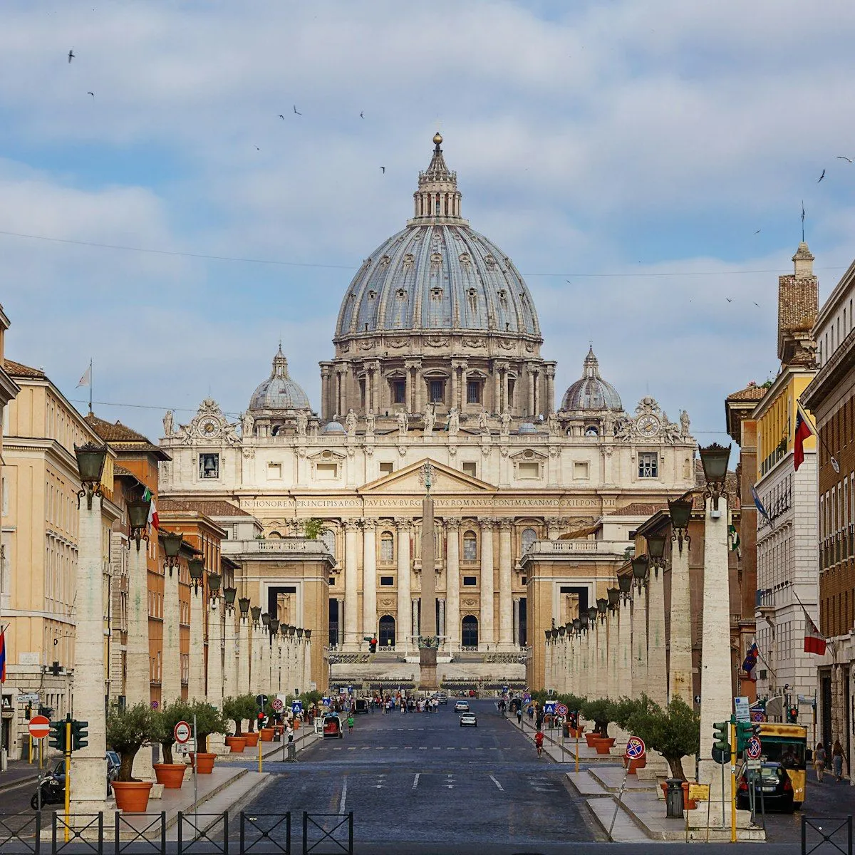 The image is a high-quality editorial shot of St. Peter's Basilica, showcasing excellent composition with the building centrally framed by the street and surrounding structures. Lighting is generally very good, providing clear detail across the scene, and colors are accurate and natural. The image is exceptionally sharp, with both the foreground and the primary architectural subject in clear focus. Its photo-realistic quality is outstanding, making it a strong representation of the landmark. The presence of some distant people and vehicles on the street is natural for the setting and does not detract from the main subject.