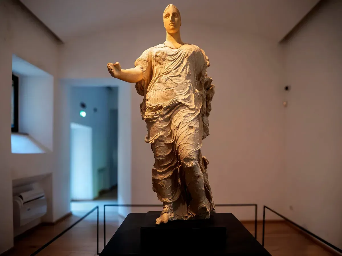 A full-length photograph of a classical-style statue, possibly depicting a draped goddess or significant figure, standing on a dark, minimalist pedestal in a museum or gallery setting. The statue is made of light-colored stone and exhibits detailed drapery.