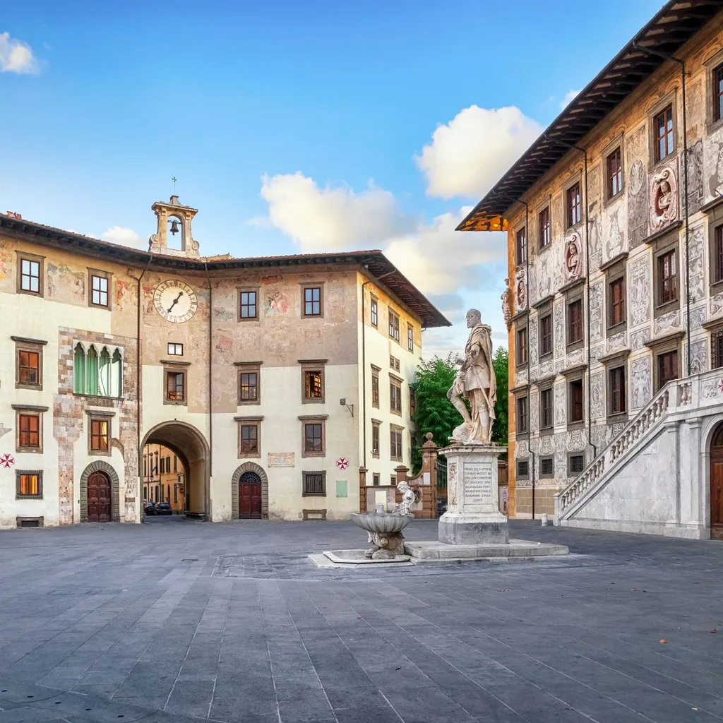 An outdoor shot of a historic European square featuring ornate stone buildings, a prominent statue, and a clear blue sky with some clouds. The square is paved with dark grey stones, and there is a fountain near the statue. The architecture includes arches, detailed facades, and multiple windows.
