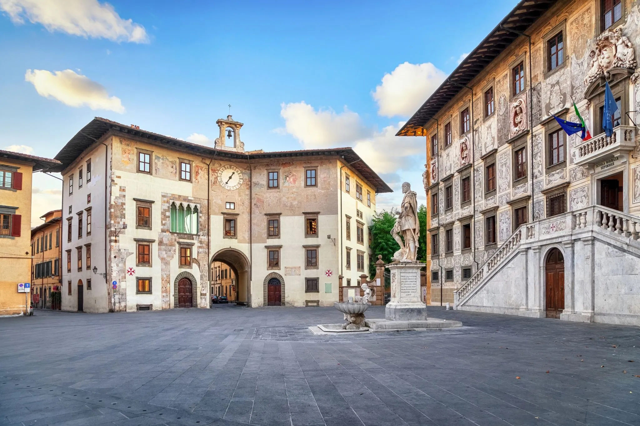 An outdoor shot of a historic European square featuring ornate stone buildings, a prominent statue, and a clear blue sky with some clouds. The square is paved with dark grey stones, and there is a fountain near the statue. The architecture includes arches, detailed facades, and multiple windows.