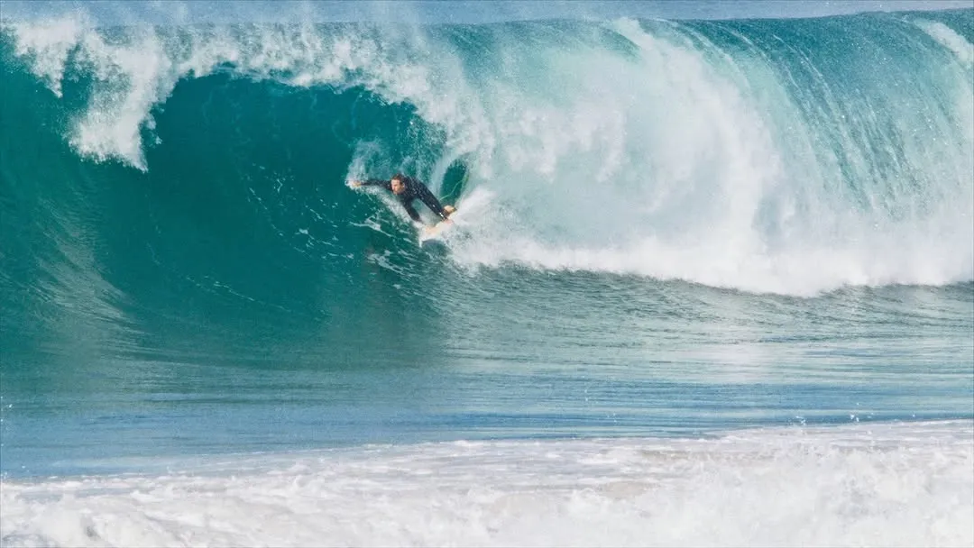 An action shot of a surfer carving through a large, powerful barrel wave. The image captures the dynamic energy of the ocean and the surfer's expertise.