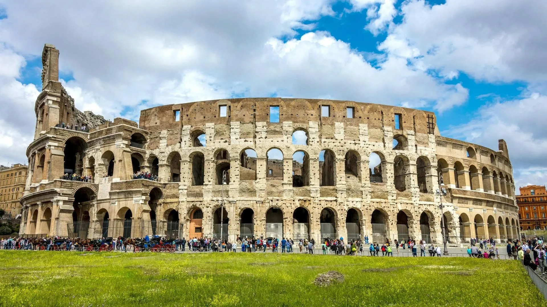 An iconic view of the ancient Roman Colosseum, captured on a bright day with a blue sky and scattered clouds overhead, and green grass in the foreground. People and horse carriages are visible near the base of the structure.