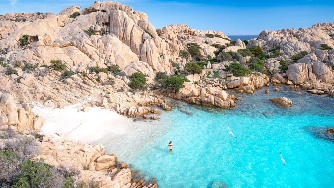 An aerial view of a stunning natural bay featuring crystal clear turquoise water, white sandy beaches, and unique light-colored rock formations under a clear blue sky. A person is seen standing in the shallow water, and a seagull flies overhead.
