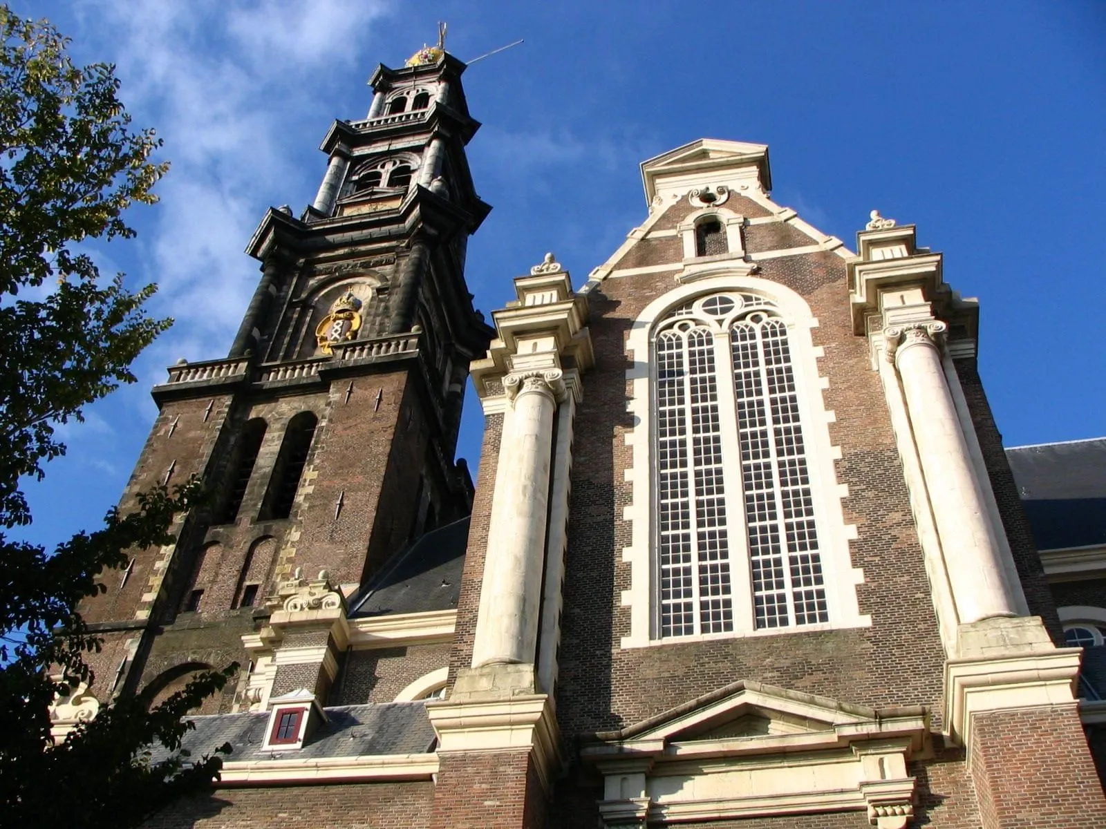 An architectural shot of a historic brick building, likely a church, featuring a tall tower and a main facade with a large arched window, all set against a bright blue sky.