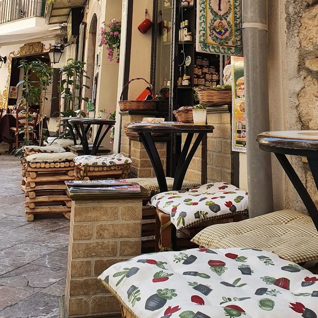 An inviting editorial-style image capturing a narrow European street scene with an outdoor cafe or shop. The image features several wooden tables and chairs with patterned cushions, leading towards a storefront adorned with hanging plants and a decorative banner. The stone architecture and climbing greenery add to the charming ambiance.