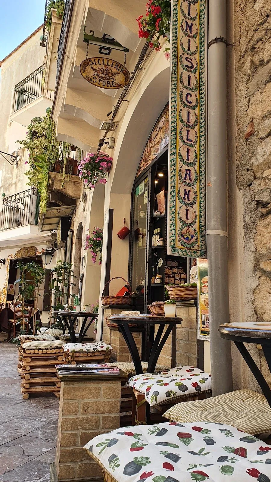 An inviting editorial-style image capturing a narrow European street scene with an outdoor cafe or shop. The image features several wooden tables and chairs with patterned cushions, leading towards a storefront adorned with hanging plants and a decorative banner. The stone architecture and climbing greenery add to the charming ambiance.