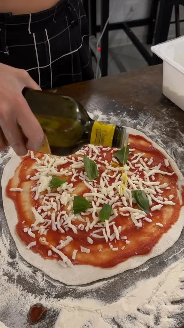 A close-up shot of a pizza being prepared, showing dough, tomato sauce, shredded cheese, basil leaves, and olive oil being drizzled from a bottle. A hand is holding the bottle.