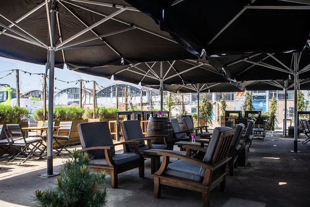 An inviting outdoor seating area featuring dark umbrellas providing shade over wooden chairs with cushions and various tables. String lights are visible in the background, adding to the ambiance. The setting appears to be a patio or urban garden space, with hints of greenery and distant buildings.