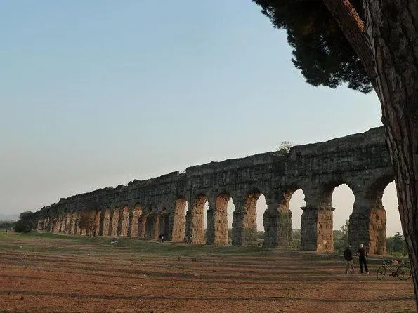 An exterior shot of the ancient Roman aqueduct, Aqua Claudia, stretching across a wide, flat landscape under a clear sky. The massive stone arches of the aqueduct show signs of age and wear. A few small figures of people are visible near the base of the structure, providing a sense of scale. A tree branch is partially visible in the upper right corner of the frame.