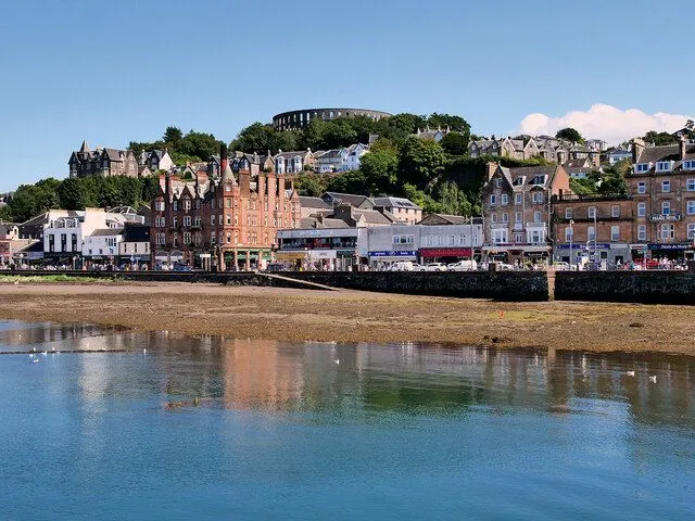 A well-composed and naturally lit photograph of a coastal town, featuring distinct layers of water, shoreline, waterfront buildings, and a hill with more structures under a clear blue sky.