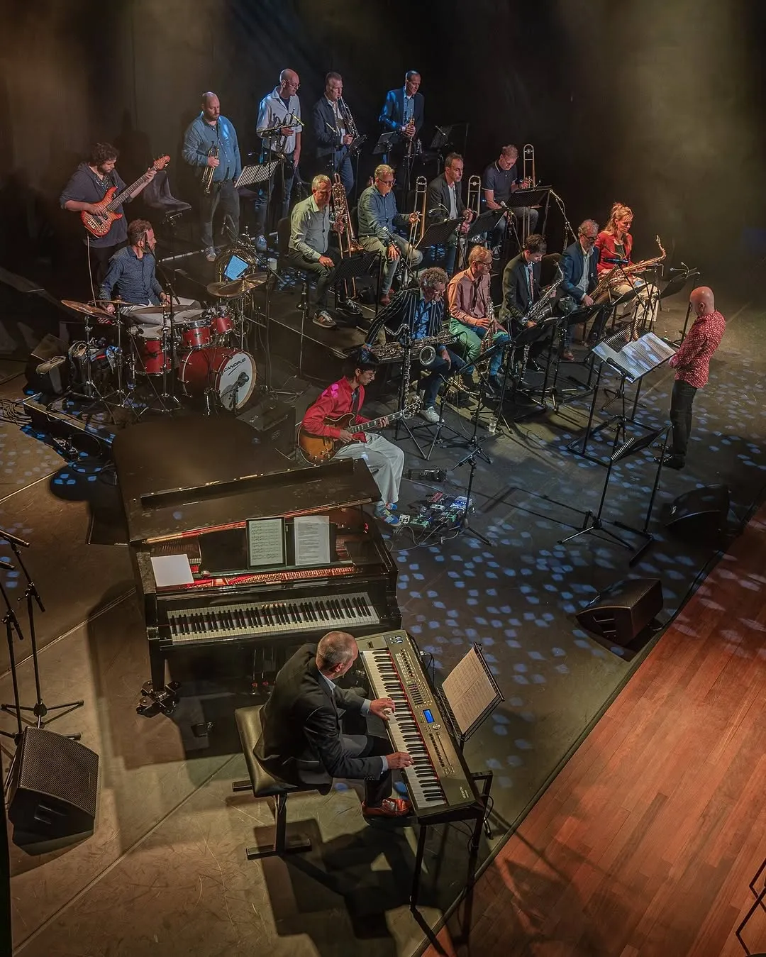 The image captures a well-composed scene of a jazz or big band performing on stage from an elevated perspective. The composition is strong, showcasing the arrangement of musicians and instruments effectively. The lighting creates a dramatic atmosphere, characteristic of a live performance, though it results in some underexposed areas. The image is photo-realistic and generally sharp, with key elements in focus. Colors appear accurate given the stage lighting conditions.