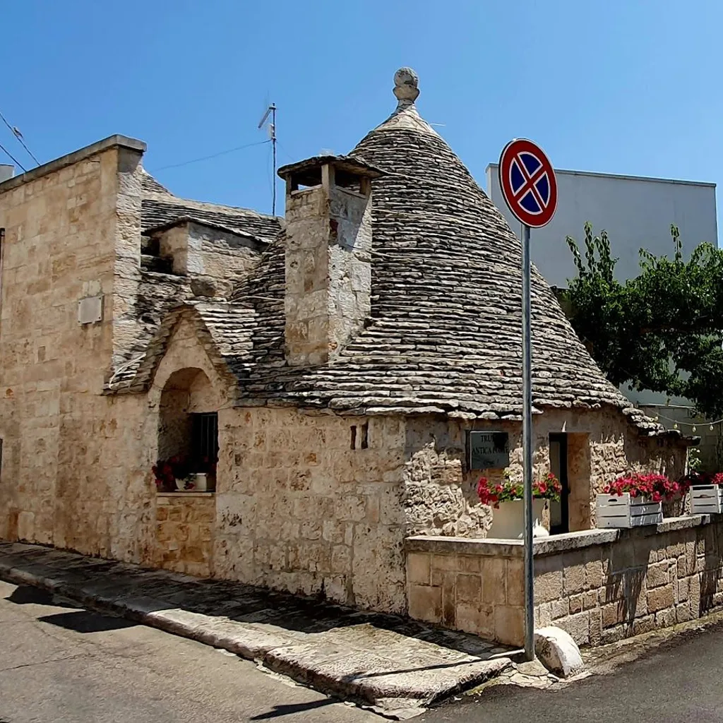An exterior view of a traditional Trullo house with its distinctive conical stone roof, set on a sunny street with a stone wall adorned with red flowers.