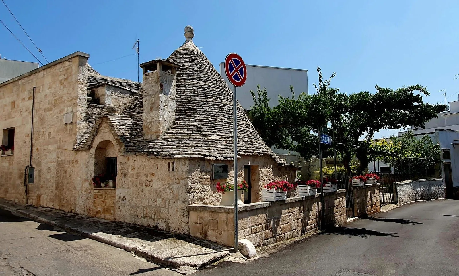 An exterior view of a traditional Trullo house with its distinctive conical stone roof, set on a sunny street with a stone wall adorned with red flowers.