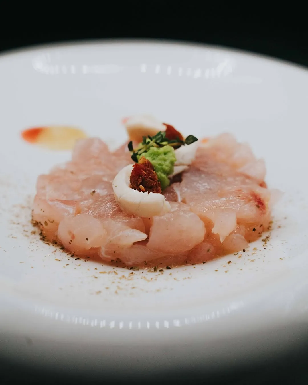 A close-up, top-down shot of a beautifully plated seafood dish, possibly a tartare or ceviche, on a white ceramic plate. The dish features finely chopped light-pink fish, garnished with green paste (possibly wasabi or avocado), white curls (possibly squid or thinly sliced onion), and small pieces of dried red ingredients (possibly sundried tomatoes). There are also spices sprinkled around the base of the dish on the plate.