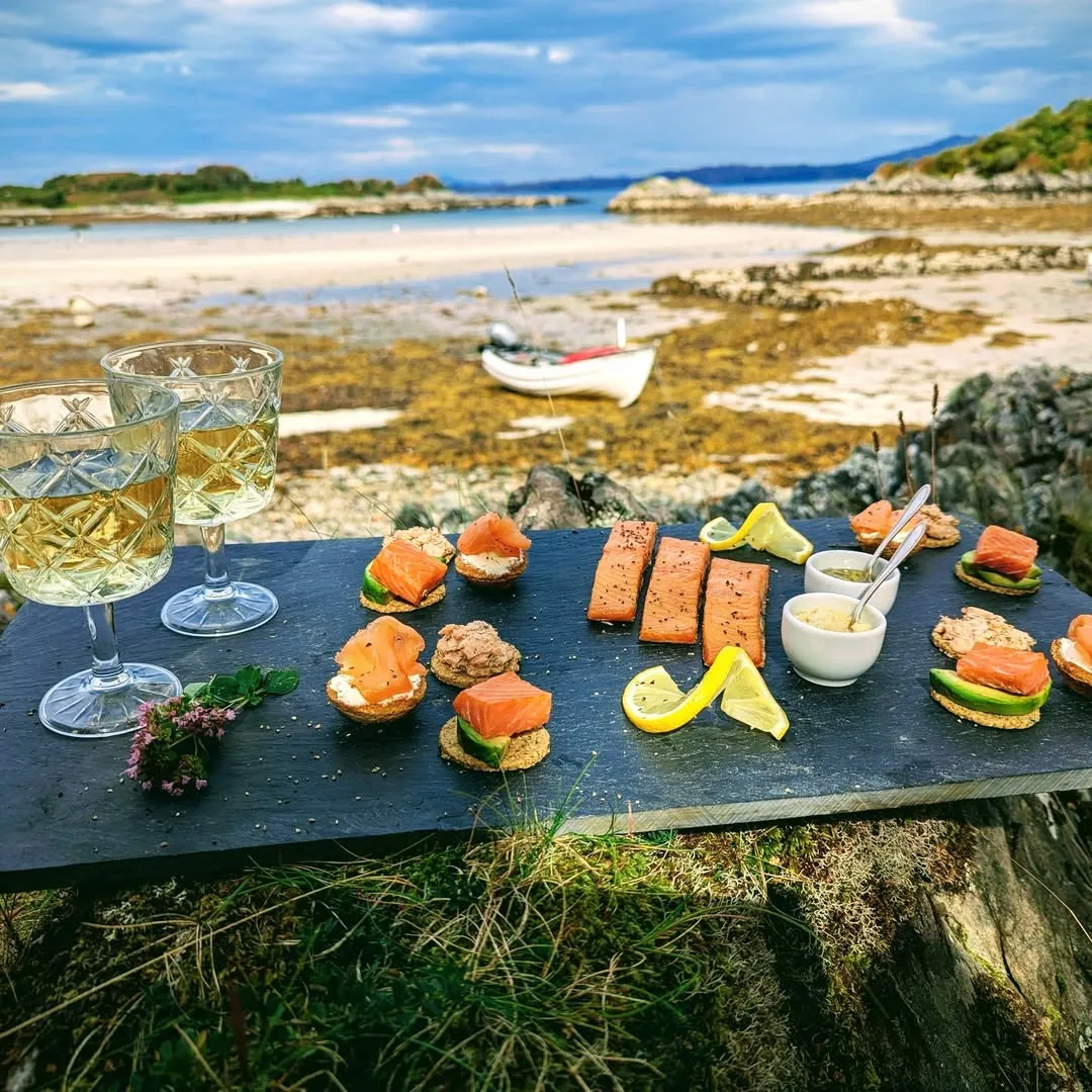 A beautifully composed shot of an outdoor picnic or dining experience on a beach, featuring a slate board laden with an assortment of canapés, likely salmon-based, and two glasses of white wine. The natural setting with the sea and sky creates a serene backdrop.
