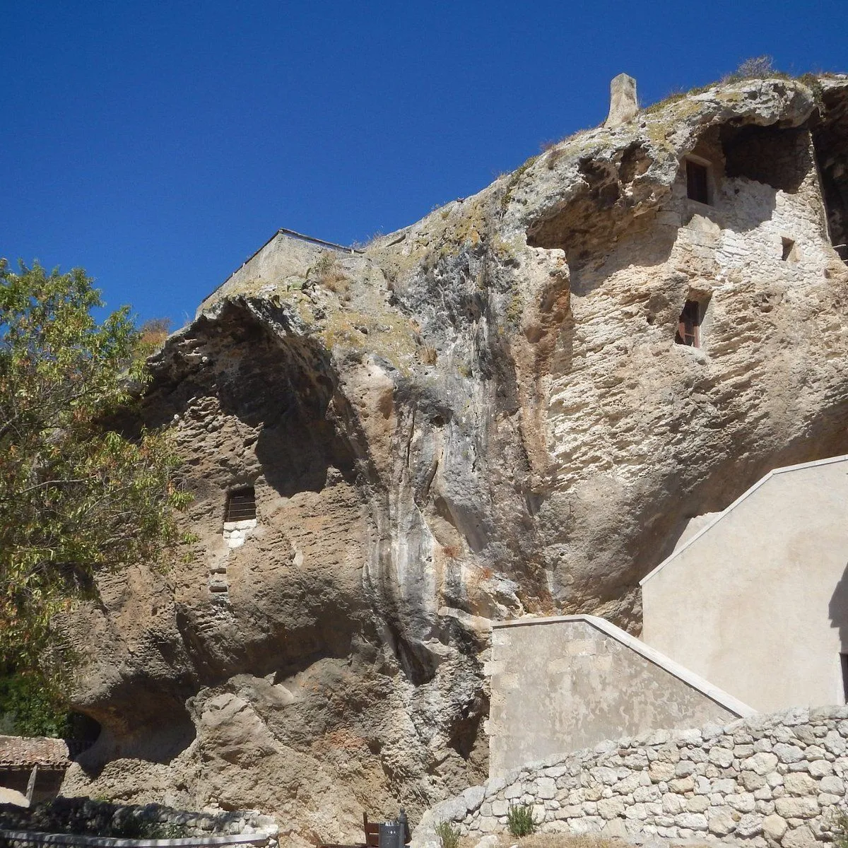 An ancient dwelling or structure carved into or built against a large rock formation under a clear blue sky. The image features natural rock textures, integrated architectural elements like windows and walls, and foreground elements such as a stone wall and stairs.