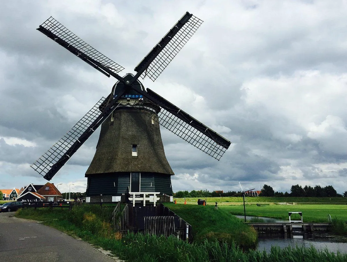 A well-composed photograph of a traditional Dutch windmill set against an overcast sky. The windmill, surrounded by lush green fields, a small canal, and a road, is the central focus. The image effectively captures the rural scenic beauty.