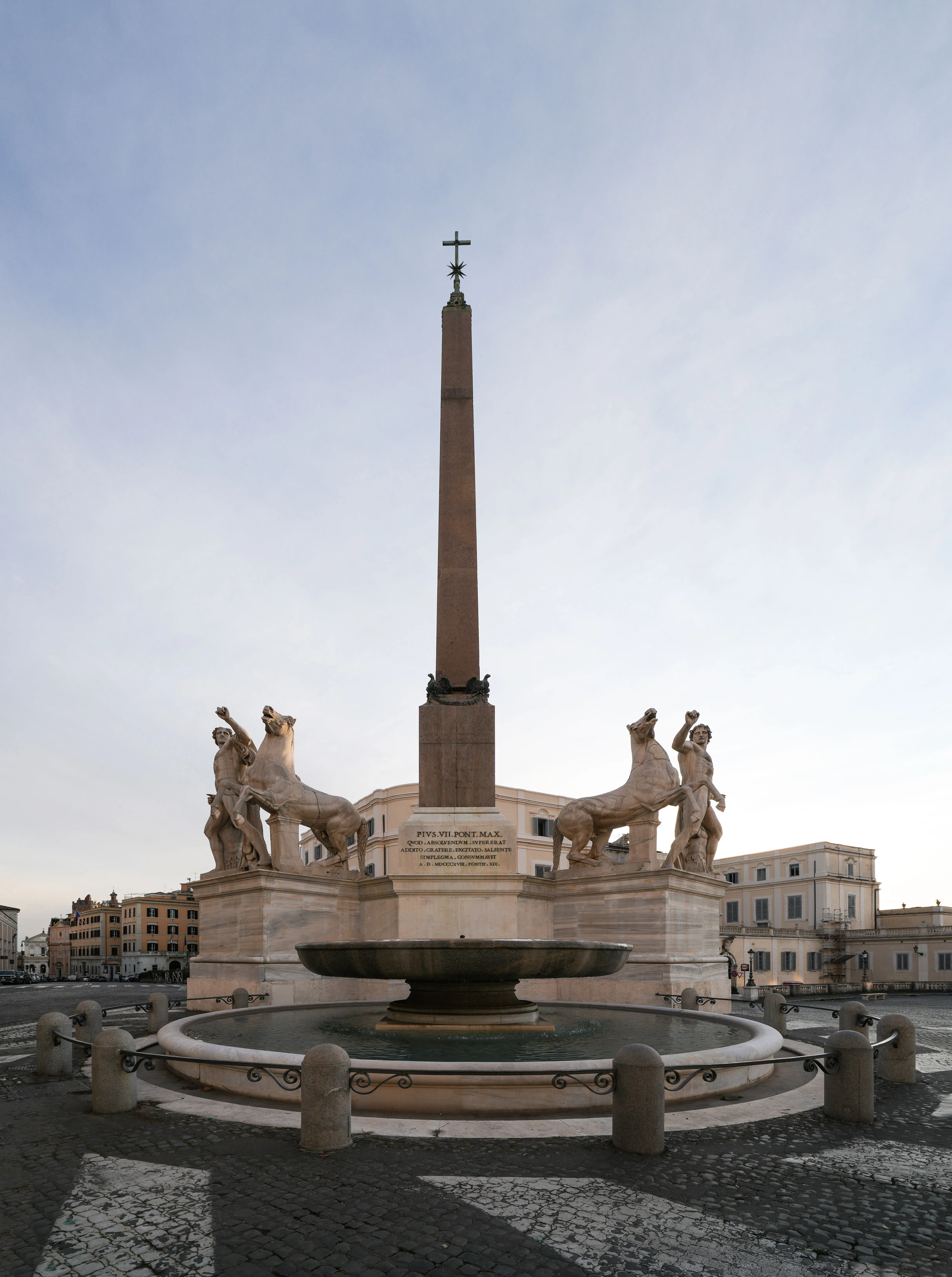 An editorial-style image of a grand historical monument featuring an obelisk, statues, and a fountain in a public square. The composition is well-balanced and symmetrical, highlighting the main architectural elements.