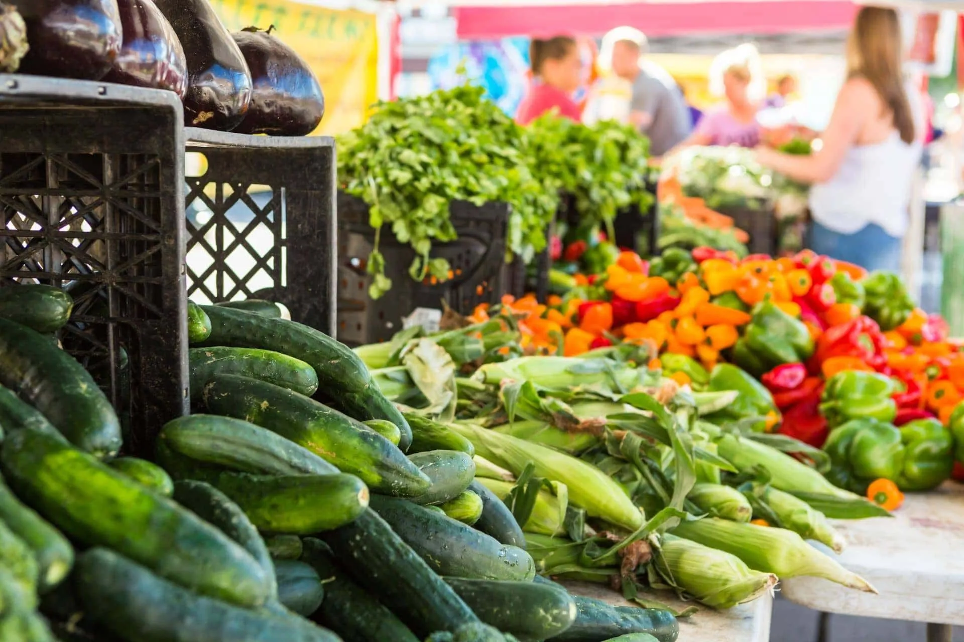 A vibrant and well-stocked produce stall at a farmer's market, featuring fresh cucumbers, corn, bell peppers, and herbs. The background shows blurred market-goers, adding context to the scene.
