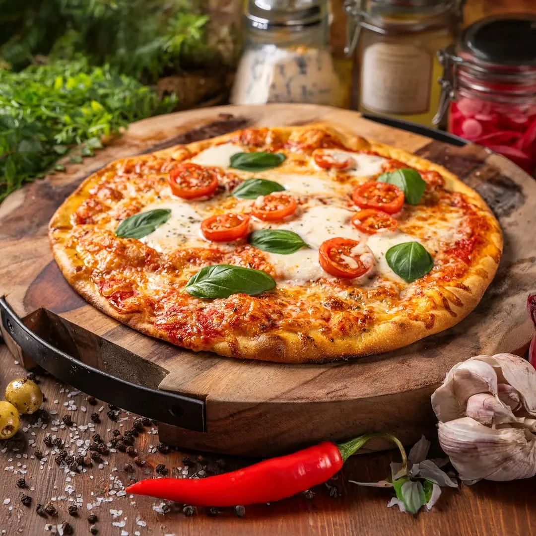 This is a high-quality, professionally shot image of a pizza. The composition is excellent, with the pizza centered on a rustic wooden board, complemented by relevant background and foreground elements like fresh herbs, spices, chili, and garlic that enhance the theme without cluttering the scene. The lighting is soft and even, highlighting the textures of the melted cheese, crisp crust, and fresh toppings. The image is exceptionally sharp, with the key elements (the pizza) in perfect focus, and the colors are vibrant and accurate, contributing to a highly photo-realistic and appealing presentation.