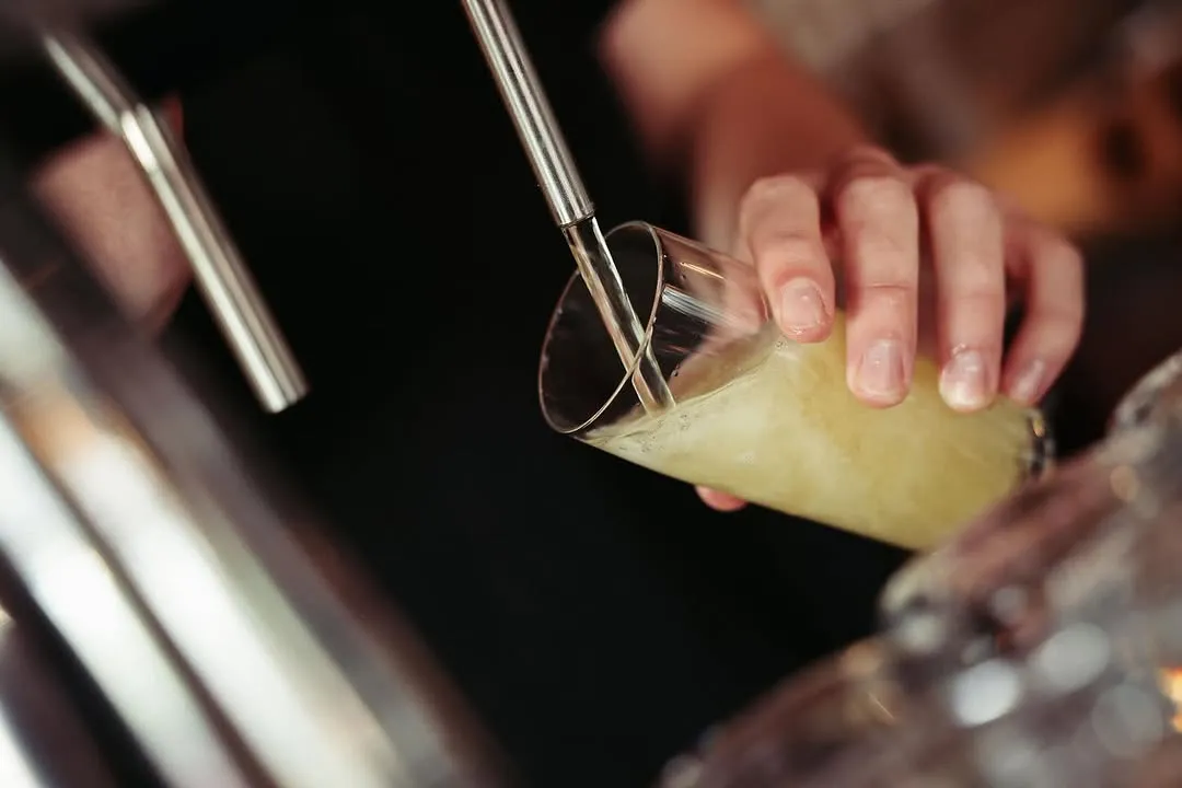 The image features a close-up of a hand holding a glass, into which a golden liquid is being poured from a tap or dispenser. The focus is sharp on the action, highlighting the liquid stream and the glass. The lighting is effective, creating a visually appealing and realistic shot with accurate colors.